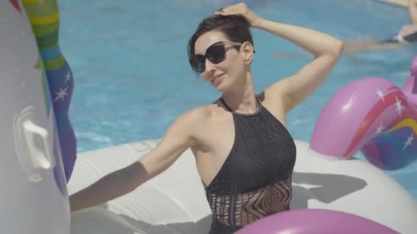 선글라스를 쓴 브루넷 여성은 수영링 위에 앉아 카메라 앞에서 웃고 있었다. 야외에서 화창 한 여름날 수영장의 호화 로운 휴양지에서 일광욕을 즐기는 백인 여성의 모습, — 비디오