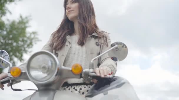 若い白人女性のオートバイのハンドルを握って座っている。スクーターの屋外で美しい笑顔の女性バイカーの肖像画。ハッピーブルネット女性楽しんで曇り夏の日. — ストック動画