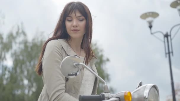 Ritratto di bella giovane donna che guarda lo specchio retrovisore della moto e ammira il riflesso. Positivo affascinante ragazza caucasica seduta su scooter sulla nuvolosa giornata estiva all'aperto. — Video Stock