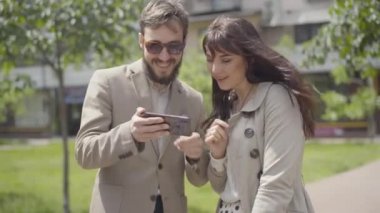 Akıllı telefonlu arkadaşlarıyla gülümseyen beyaz adam ve kadın. Mutlu yetişkin insan grubunun portresi güneşli yaz kenti caddesinde buluşuyor..