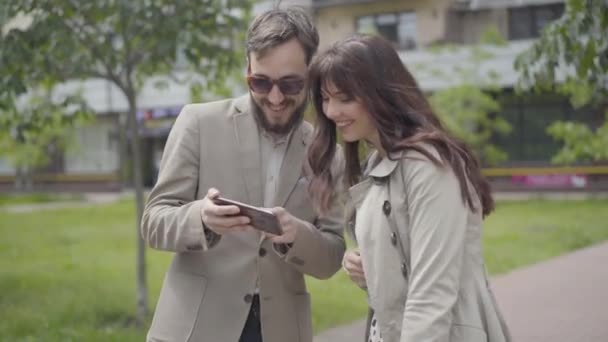 Positive Caucasian couple surfing Internet in smartphone outdoors. Portrait of smiling young man and woman using social media standing on city street and talking. — Stock Video