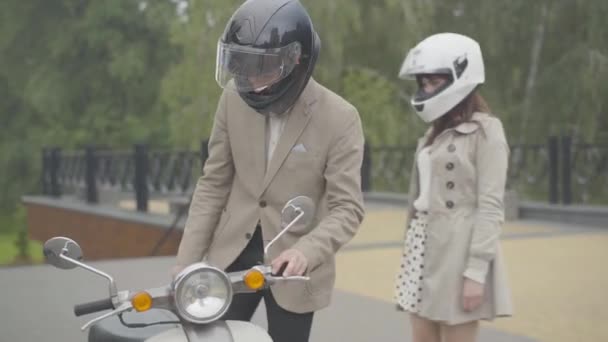 Couple de motards caucasiens dans les casques venant à scooter à l'extérieur et assis sur elle. Homme et femme élégants confiants jouissant de passe-temps le jour de l'été. — Video