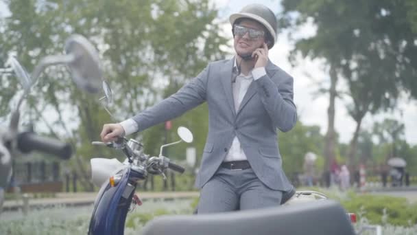 Retrato de un hombre seguro y positivo en casco y gafas de sol hablando por teléfono. Conductor de scooter caucásico sonriente en traje de negocios formal sentado en bicicleta al aire libre en el día nublado de verano. — Vídeos de Stock