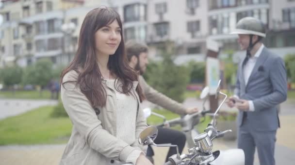 Pewna siebie piękna kobieta siedząca na retro hulajnodze trzymająca kask w dłoniach jako dwóch zamazanych mężczyzn mówiących w tle. Portret pozytywnych białoruskich motocyklistów odpoczywających na ulicy miejskiej. — Wideo stockowe