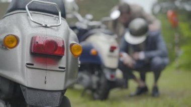 Arka planda motorsikleti tamir eden iki bulanık adamın yanıp söndüğü yakın çekim. Motorcular yağmurlu bir yaz gününde açık havada trafik kazası geçiriyorlar..