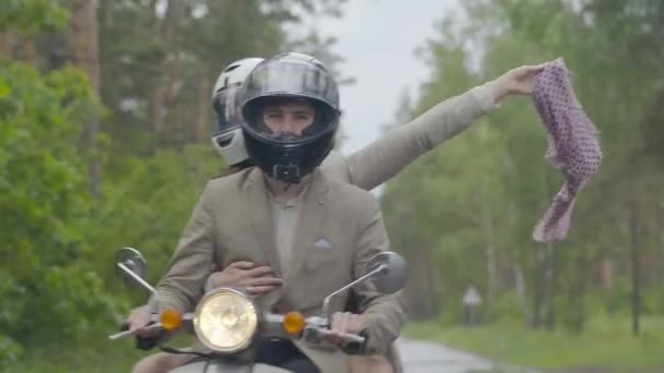 Femme heureuse assise sur le siège arrière de la moto avec les mains s'étire tenant foulard. Homme souriant scooter de conduite avec petite amie à l'arrière. Couple adulte positif sur la route le jour nuageux. — Video