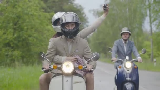 Alegre grupo de amigos montando scooters y grabando video de un viaje. Hombres caucásicos confiados conduciendo motocicletas como mujer sentada en el asiento trasero con cámara y disparos. — Vídeos de Stock