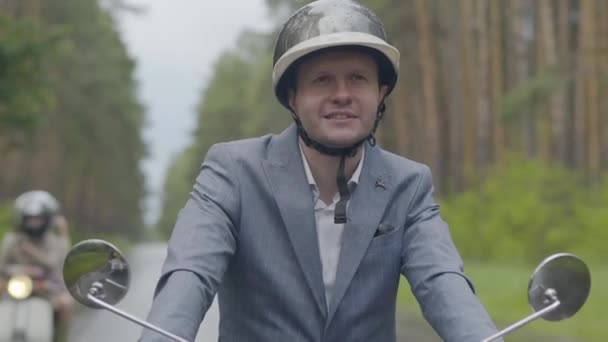 Retrato de cerca del feliz hombre sonriente conduciendo motocicleta al aire libre. Alegre ciclista adulto caucásico en casco a caballo en scooter en la carretera suburbana en el día nublado de verano. — Vídeos de Stock