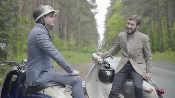 Dos hombres caucásicos seguros y positivos sentados en scooters retro en el campo y hablando. Retrato de elegantes motociclistas en trajes formales charlando y riendo como descansando en viaje por carretera. — Vídeos de Stock