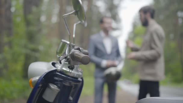 Крупный план мотоциклетного руля с двумя размытыми людьми, разговаривающими на заднем плане. Ретро скутер стоит на улице в пасмурный день в сельской местности, пока друзья-мужчины болтают. — стоковое видео