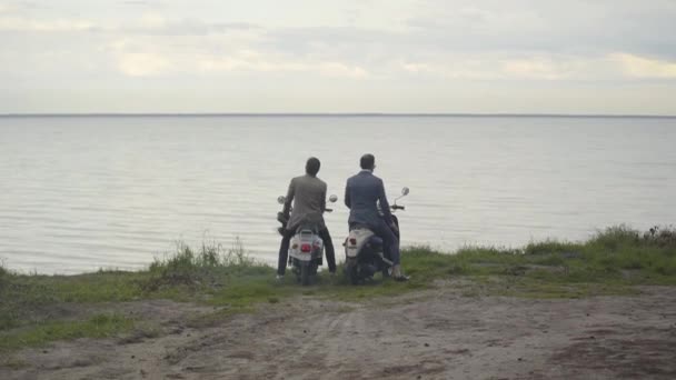 Extreme wide shot of two confident elegant men getting off scooters and looking at picturesque waterscape. Back view of male Caucasian friends in formal suits and helmets resting on river bank. — Stock Video