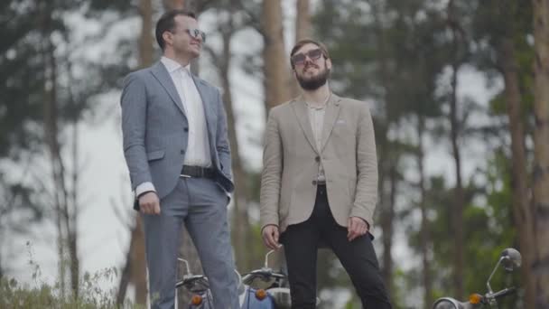 Retrato de dos hombres caucásicos positivos en gafas de sol de pie en el bosque y hablando. Felices ciclistas masculinos disfrutando de un viaje de verano al aire libre en un día nublado. — Vídeos de Stock