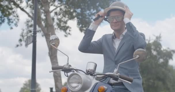 Retrato de plano medio del hombre seguro positivo que se pone el casco en slow mo como sentado en la motocicleta al aire libre. Motociclista caucásico sonriente antes de viajar en el día lluvioso de verano. Sede del cine 4k ProRes. — Vídeo de stock