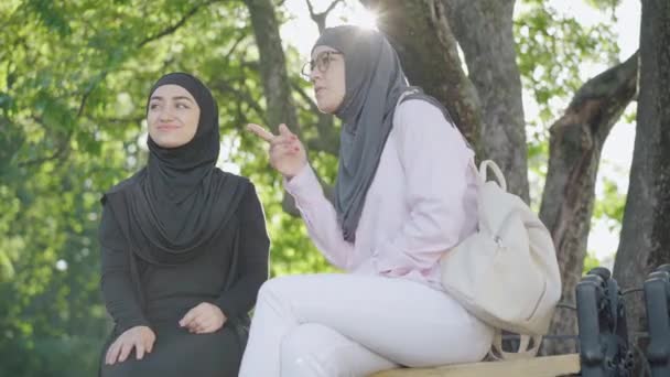 戴着眼镜和头巾的聪明的穆斯林妇女的侧面视图，她指着一边，与微笑的朋友交谈。在室外公园里阳光灿烂的日子里，积极的女性形象得到了体现。休闲概念. — 图库视频影像