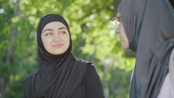 Retrato de close-up de bela jovem muçulmana em hijab preto conversando com amigo borrado e sorrindo. Encantadora senhora confiante conversando com o grupo feminino ao ar livre no dia ensolarado de verão . — Vídeo de Stock
