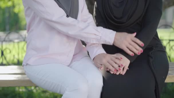 La cámara se mueve hacia arriba desde las manos femeninas abrazándose unas a otras a las caras tristes de dos mujeres musulmanas sentadas en el soleado parque al aire libre. Mujeres inmigrantes molestas que tienen problemas. Concepto de desafíos. — Vídeo de stock