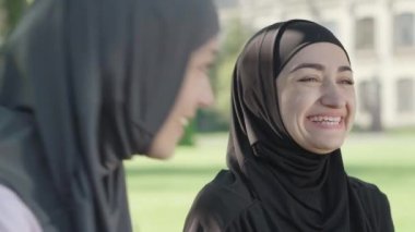 Siyah geleneksel tesettürlü gülen Müslüman kadının açık havada güneşli bir günün tadını çıkardığı yakın plan. Üniversite bahçesinde arkadaşlarıyla dinlenen neşeli genç bayan portresi. Mutluluk kavramı.