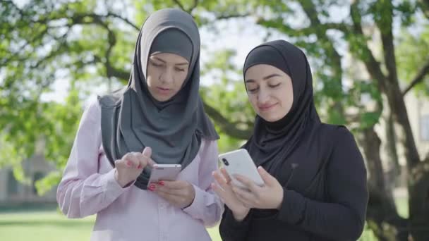 Mulheres muçulmanas modernas confiantes usando smartphones ao ar livre. Retrato de duas jovens belas senhoras em hijabs surf Internet no dia ensolarado de verão ao ar livre. Estilo de vida, confiança . — Vídeo de Stock