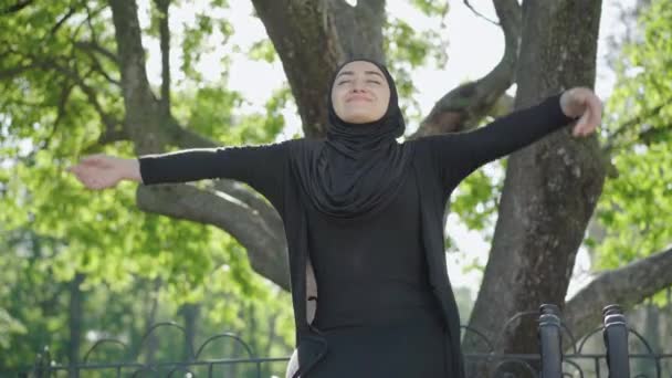 Retrato de mulher muçulmana jovem feliz em roupas tradicionais pretas e hijab desfrutando de dia ensolarado de verão no parque. Sorrindo encantadora imigrante feminino descansando ao ar livre. Conceito de liberdade e estilo de vida . — Vídeo de Stock