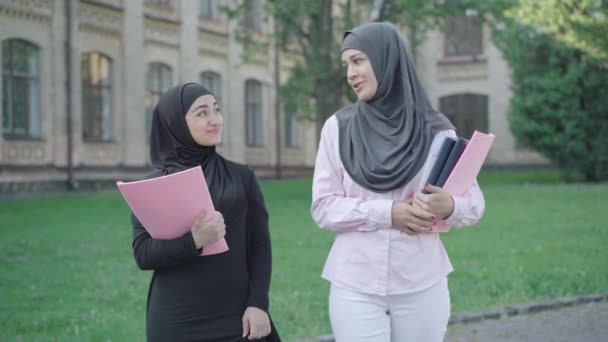Retrato de duas mulheres muçulmanas confiantes andando no pátio da universidade e conversando. Jovens imigrantes do sexo feminino a estudar na Europa. Modernas senhoras do Oriente Médio desfrutando de educação no exterior . — Vídeo de Stock