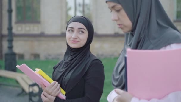 Oldalnézetben magabiztos muszlim diákok sétálnak az egyetemi udvaron és beszélgetnek. Két pozitív, gyönyörű nő sétál ki a szabadba a napsütéses napon. Külföldön tanuló bevándorlók Európában. Oktatási koncepció. — Stock videók