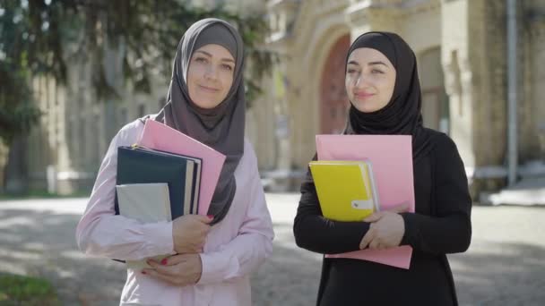 Feliz sorrindo mulheres muçulmanas posando no quintal da universidade no dia ensolarado de verão. Retrato de duas alunas confiantes em pé na frente da faculdade ao ar livre. Estilo de vida moderno e conceito de educação . — Vídeo de Stock