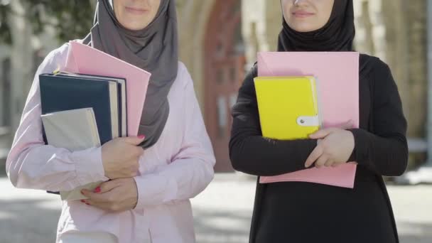 Two unrecognizable Muslim student posing outdoors with book. Front view portrait of confident positive ladies in traditional hijabs standing at university yard on sunny day and smiling at camera. — Stock Video