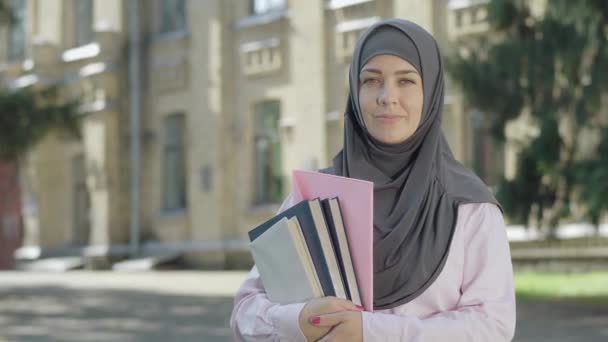 Portret van een gelukkige moslimvrouw in grijze hijab poserend voor de universiteit op zonnige dag. Positieve vrouwelijke immigrant studeert aan de Europese universiteit. Vrolijke jongedame met boeken die glimlachen op de camera. — Stockvideo