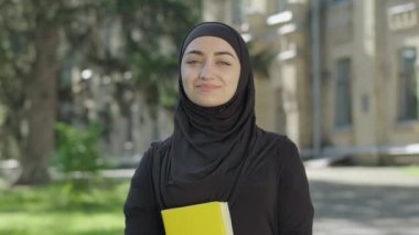 Güneşli bir günde üniversite bahçesinde poz veren geleneksel siyah tesettürlü, gururlu Müslüman bir kadın. Kitapları tutan ve kameraya gülümseyen kendinden emin kız öğrencinin portresi. Güven ve eğitim.