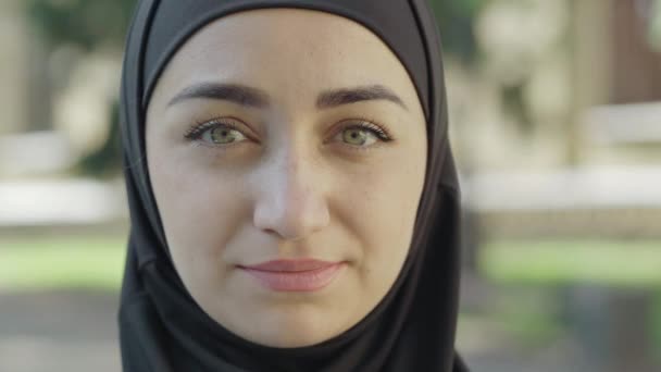 Close-up rosto de jovem mulher bonita em hijab tradicional olhando para a câmera e sorrindo. Retrato de senhora muçulmana encantadora confiante posando ao ar livre no dia ensolarado. Conceito de diversidade e beleza . — Vídeo de Stock