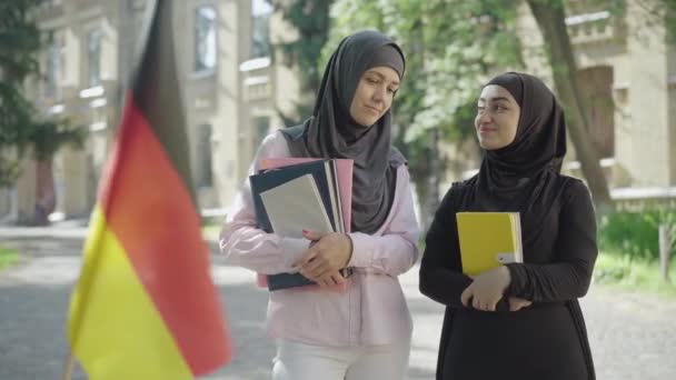大学の庭に本を持って立っている2人の女性イスラム教徒の移民と正面になびくぼやけたドイツの旗のように話しています。ヨーロッパの大学で学ぶ幸せな若い女性の肖像画. — ストック動画