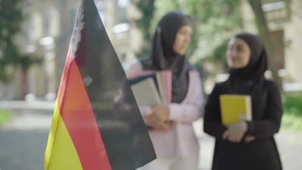 Primer plano de la bandera alemana ondeando al aire libre con dos mujeres musulmanas hablando al fondo. Mujeres inmigrantes felices estudiando en Europa. Educación internacional, concepto de inteligencia. — Vídeos de Stock