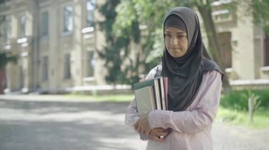 Ellerinde kitap tutan ve dışarıda bekleyen üzgün, genç Müslüman kadın. Güneşli bir günde ayrılan tesettürlü güzel üzgün üniversite öğrencisinin portresi. Üniversiteye kabul konsepti.