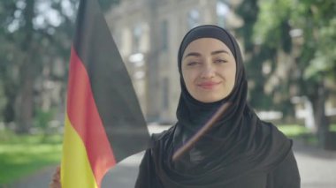 Güneşli üniversite bahçesinde Alman bayrağıyla poz veren mutlu gülümseyen Müslüman kadın portresi. Almanya 'da kolejde okuyan geleneksel siyah tesettürlü kendinden emin genç bir öğrenci. Eğitim ve zeka.