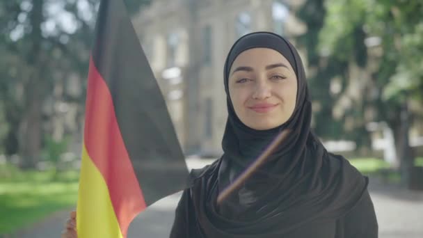 Portrét šťastně se usmívající muslimky pózující s německou vlajkou na slunném univerzitním dvoře. Důvěryhodný mladý student v tradičním černém hidžábu studující na vysoké škole v Německu. Vzdělávání a inteligence. — Stock video