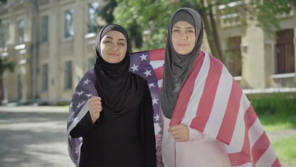Dwie muzułmanki owinięte w amerykańską flagę, patrzące w kamerę i uśmiechnięte. Portret pewnej siebie imigrantki pozującej na podwórku uniwersyteckim w USA. Happy groupmates studiuje w Stanach Zjednoczonych. — Wideo stockowe
