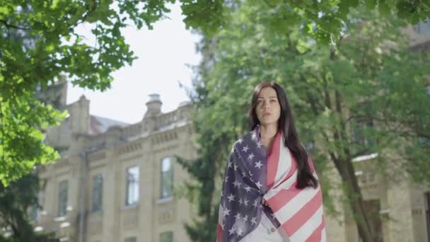 Schöne brünette Frau in amerikanische Flagge eingehüllt hebt die Hand. Porträt einer selbstbewussten, charmanten Dame, die auf dem College-Hof Freiheitsgeste zeigt. Junge Studentin steht an sonnigem Tag im Freien. — Stockvideo