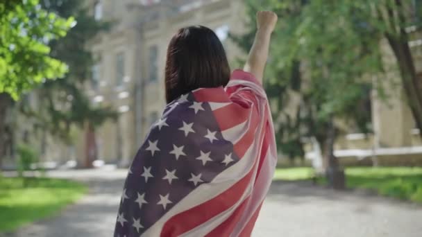Πίσω όψη της μελαχρινής γυναίκας με την αμερικάνικη σημαία να κάνει χειρονομία με το χέρι. Νεαρή λεπτή κυρία που δείχνει σημάδια ελευθερίας στην αυλή του πανεπιστημίου. Έννοια της ανεξαρτησίας ή της διαμαρτυρίας. — Αρχείο Βίντεο