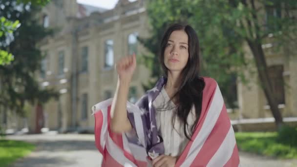 Retrato de una joven confiada en bandera americana haciendo gesto de la mano de la libertad en el patio de la universidad. seria hermosa morena estudiante posando para la libertad en el fondo de la universidad en día soleado. — Vídeos de Stock