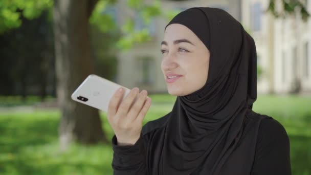 Uśmiechnięta muzułmanka nagrywająca wiadomość audio na smartfonie. Portret pewnej siebie młodej uczennicy w hidżabie korzystającej z Internetu do komunikacji bezprzewodowej siedzącej na podwórku uniwersyteckim. — Wideo stockowe
