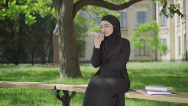 Wide shot portrait of happy Muslim woman recording audio message for wireless chat. Smiling young female student chatting online as sitting at university yard. Immigrant studying in college abroad. — Stock Video