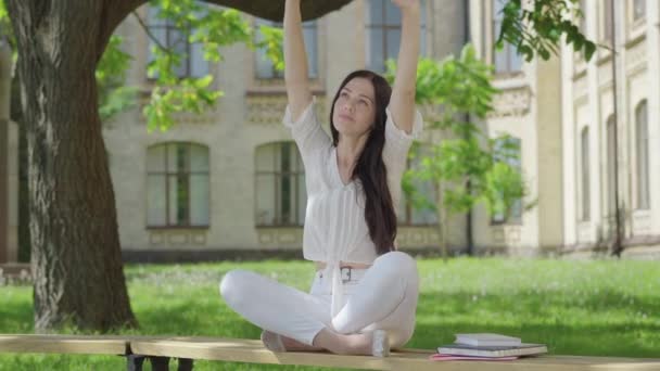 Portret van zelfverzekerde jonge vrouw in witte kleren ontspannen in lotus poseren buiten. Breed shot van mooie slanke blanke universiteitsstudent zittend op bank op zonnige dag en mediterend. Yoga-concept. — Stockvideo