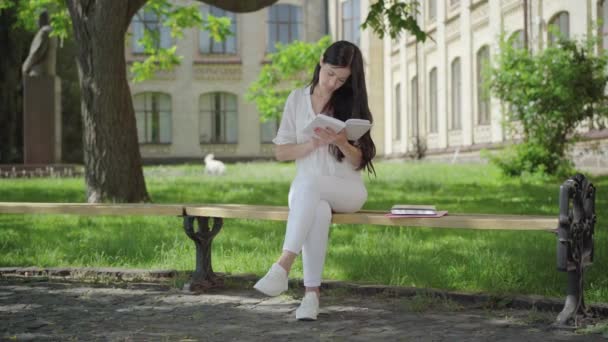 Široký záběr soustředěné brunetky v bílých šatech, jak sedí na lavičce za slunečného dne a čte si. Portrét inteligentního sebevědomého vysokoškolského studenta, který si užívá koníček venku. Koncept vzdělávání. — Stock video