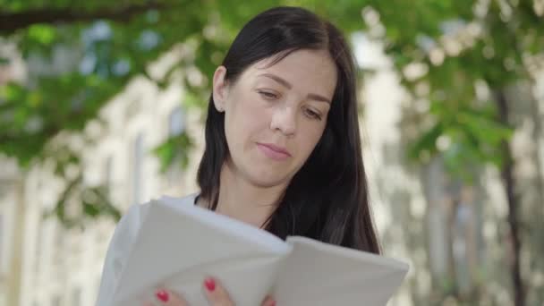 Cara de cerca de una mujer caucásica absorta leyendo un libro en el patio de la universidad. Retrato de una morena concentrada disfrutando de la literatura al aire libre en un día soleado. Hobby y concepto de estilo de vida. — Vídeos de Stock