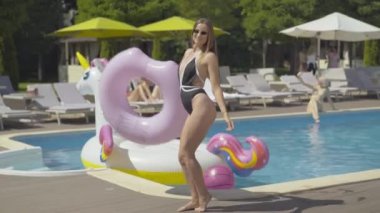 Kendine güvenen zayıf bir kadının havuz kenarında donut yüzme yüzüğüyle dans ettiği geniş açı. Güzel esmer beyaz turistin portresi güneşli bir günde lüks yaz otelinde tatilin tadını çıkarıyor..
