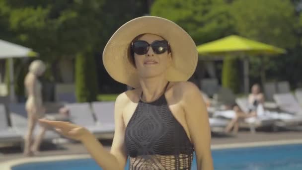 Retrato de una joven alegre con sombrero de sol y gafas de sol bailando en un lujoso resort junto a la piscina. Encantadora turista morena disfrutando de vacaciones en el soleado día de verano al aire libre. — Vídeo de stock