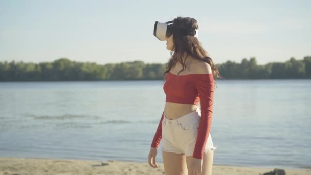 Überraschte junge Kaukasierin mit VR-Brille, die sich im Freien umschaut. Porträt einer aufgeregten brünetten Dame im Virtual-Reality-Simulator, die sich an sonnigen Tagen am Sommerstrand ausruht. — Stockvideo