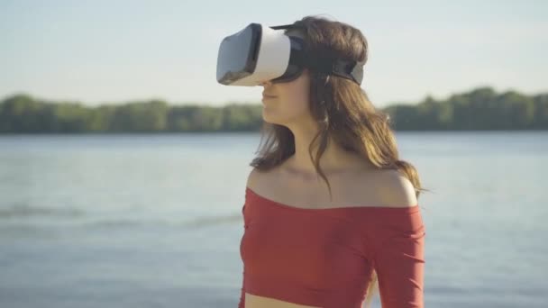 Portrait de jeune femme brune en lunettes VR regardant autour avec une expression faciale surprise excitée. Mince bronzé dame blanche debout sur la rive de la rivière dans les rayons de soleil profiter de la réalité virtuelle. — Video