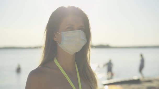 Gros plan de la belle jeune femme en masque médical debout au soleil sur la plage d'été au coucher du soleil. Portrait d'une touriste caucasienne aux rayons du soleil à la station balnéaire de Covid-19. Tourisme pandémique. — Video