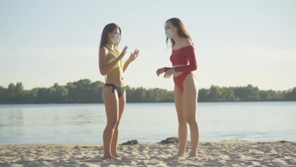 Szerokie ujęcie szczęśliwych młodych bosych turystów w strojach kąpielowych i maskach na twarzy stojących na piaszczystej plaży o zachodzie słońca i mówiących. Pozytywne kobiety rasy kaukaskiej odpoczywające na zewnątrz w kurorcie pandemicznym Covid-19. — Wideo stockowe
